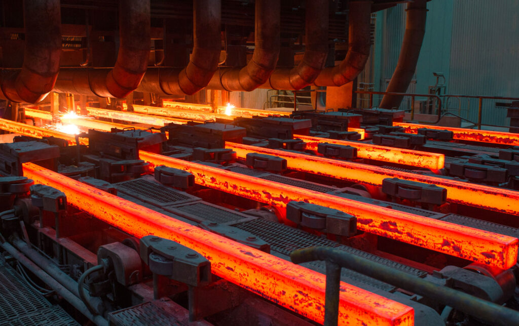Fotografia de uma siderúrgica em pleno funcionamento, mostrando barras de aço bruto superaquecidas sendo produzidas. As barras incandescentes são visíveis saindo do processo de fabricação e sendo transportadas por esteiras.