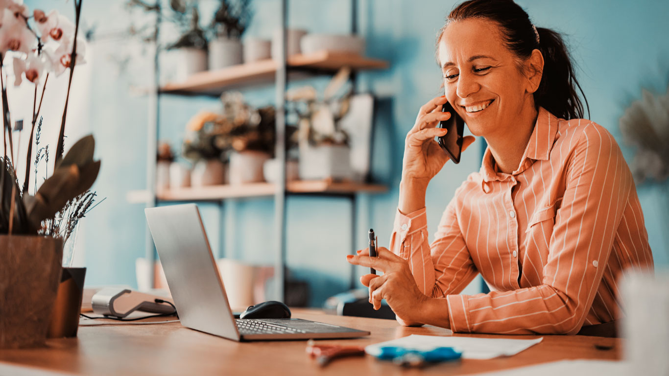 Microempresária atende o telefone e sorri. Ela está sentada numa mesa segurando uma caneta de frente para um notebook,